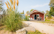 Udendørs billede af en hyggelig rød lodge med veranda, hvor to personer sidder ved et bord i solen.