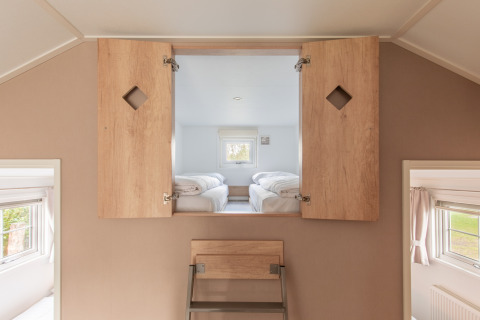 Dachbodenraum mit Leiter und offenen Holztüren, die zu einem Schlafzimmer mit zwei Einzelbetten führen.