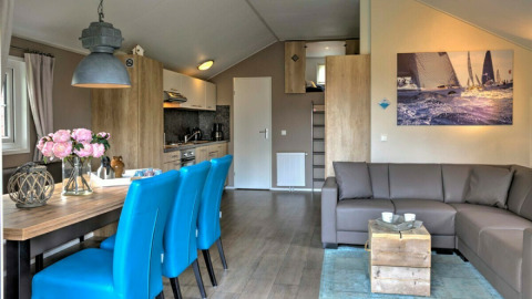 Modernes Lodge-Wohnzimmer mit blauen Stühlen, grauem Sofa, Holztisch und Küche im Vakantiepark Leukermeer.