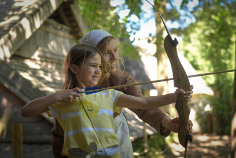 Niña y adulta practicando tiro con arco frente a una tienda safari en Lodgetent Archeon Expedition, Países Bajos.