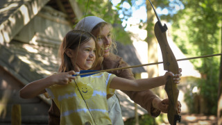 Niña y adulta practicando tiro con arco frente a una tienda safari en Lodgetent Archeon Expedition, Países Bajos.