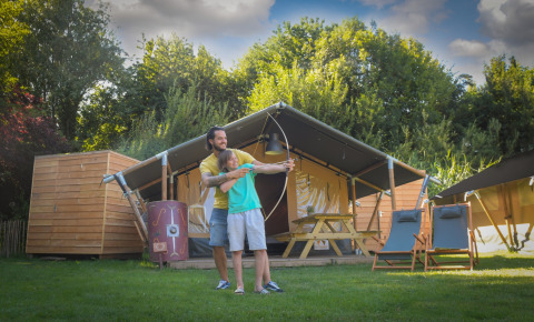 Un adulto ayuda a un niño con el tiro con arco frente a una tienda safari en Lodgetent Archeon Expedition.