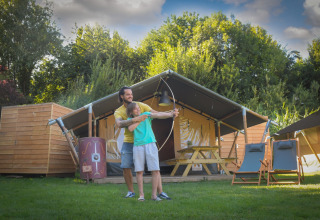 Un adulto ayuda a un niño con el tiro con arco frente a una tienda safari en Lodgetent Archeon Expedition.