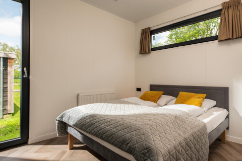 Modern bedroom at Oak Lodge with double bed, yellow pillows, large windows and view of green outdoors.