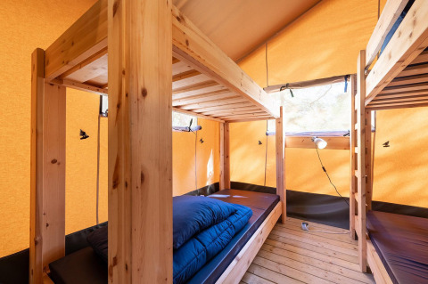 Interior de una tienda safari Villatent Wood en Camping River, Francia, con literas de madera y ropa de cama.