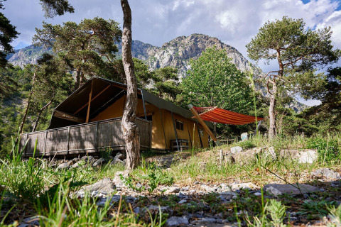 Tienda safari Villatent Wood en Camping River, Francia, rodeada de árboles, montañas y cielo azul claro.