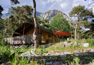 Tienda safari Villatent Wood en Camping River, Francia, rodeada de árboles, montañas y cielo azul claro.