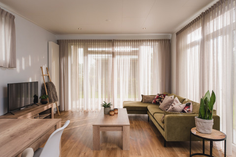 Soggiorno luminoso e moderno in un lodge di lusso al Vakantiepark Hölte, Paesi Bassi, con divano verde e grandi finestre.