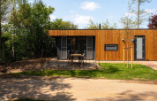 Moderne træhytte kaldet Oak Lodge ved Vakantiepark Hölte, Holland, omgivet af grøn have og træer.