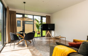 Moderne stue i Oak Lodge på Vakantiepark Hölte i Holland med store vinduer, sofa, spisebord og planter.