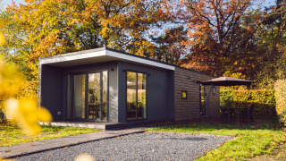 Tiny house moderne entourée d’arbres d’automne, terrasse avec parasol et meubles de jardin.