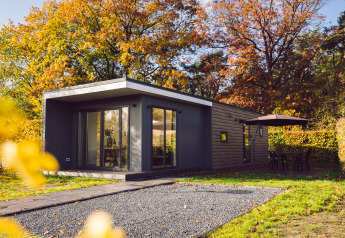 Tiny house moderne entourée d’arbres d’automne, terrasse avec parasol et meubles de jardin.