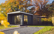 Moderne tiny house in de natuur, omringd door herfstbomen en een terras met parasol en tuinmeubilair.