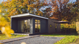 Pequeña casa moderna rodeada de árboles otoñales, con patio, sombrilla y muebles de exterior.