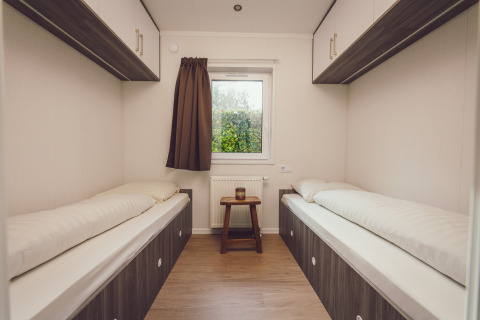 Twin single beds in a tiny house bedroom with a window, brown curtain, and wooden floor decor.