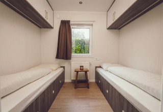 Due letti singoli in una piccola camera di tiny house con finestra, tenda marrone e pavimento in legno.