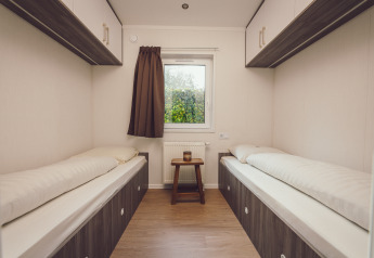 Due letti singoli in una piccola camera di tiny house con finestra, tenda marrone e pavimento in legno.