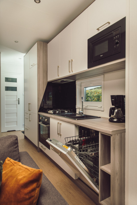 Cocina moderna de tiny house con lavavajillas abierto, horno, microondas y cafetera sobre la encimera.