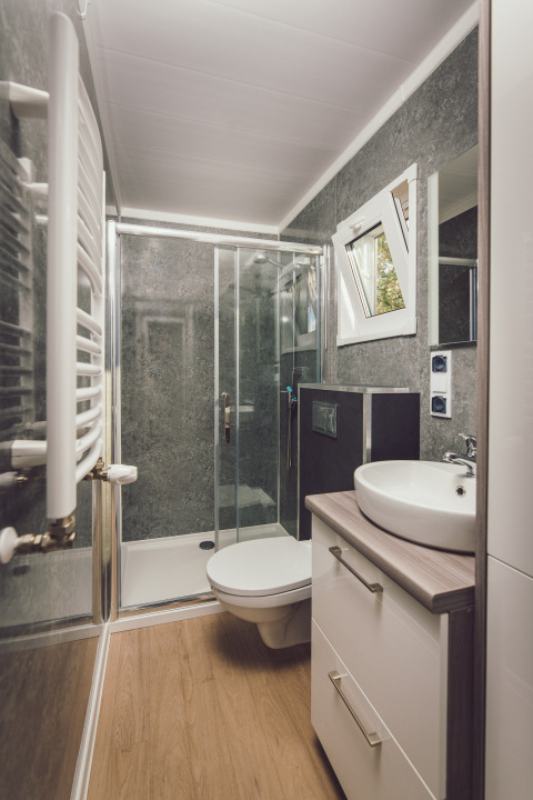 Salle de bain moderne et compacte dans une tiny house avec douche, lavabo, toilettes et sol aspect bois.