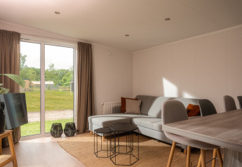 Helle Tiny-House-Wohnung mit grauem Sofa, Esstisch und Blick ins Grüne im Vakantiepark Hölte, Niederlande.