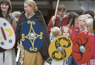 Børn og voksne iført historiske kostumer med farverige skjolde under en begivenhed i Archeon, Holland.