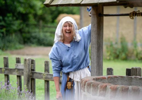 Mujer sonriente con vestimenta medieval junto a un pozo en Expedition Archeon, parque vacacional en Holanda del Sur.
