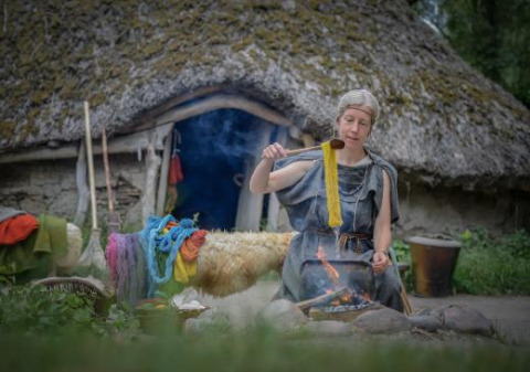 Mujer tiñendo hilo sobre fuego al aire libre frente a una casa de techo de paja en Expedition Archeon, Holanda.
