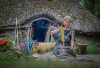 Donna che tinge filato sul fuoco davanti a una casa con tetto di paglia all’Expedition Archeon, Paesi Bassi.