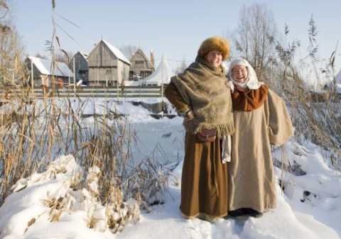 To personer i middelaldertøj står smilende i sneen nær Alphen aan den Rijn, med historiske bygninger bagved.