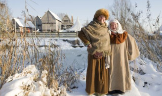 Dos personas con ropa medieval sonríen en un paisaje nevado cerca de Alphen aan den Rijn, edificios antiguos al fondo.