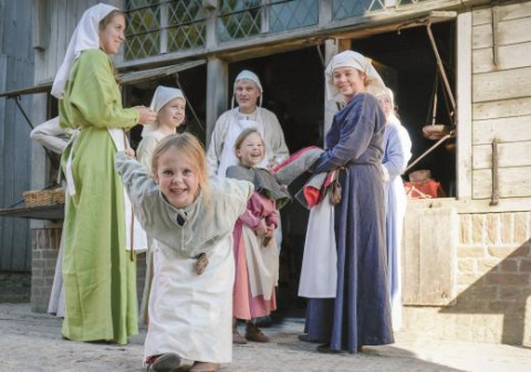 Niños y adultos con disfraces medievales juegan y sonríen afuera de una casa de madera en Expedition Archeon.