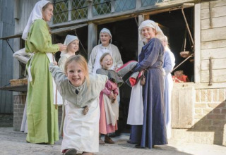 Kinderen en volwassenen in middeleeuwse kostuums spelen en lachen bij een houten gebouw in Expedition Archeon.