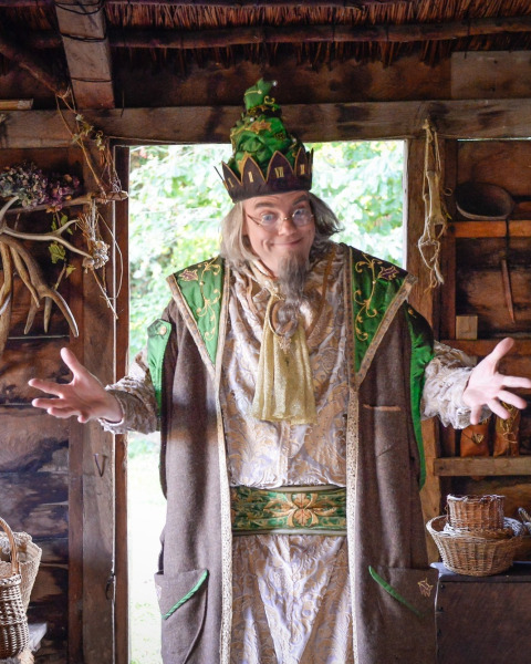 Een man in fantasierijke kostuums en kroon poseert met open armen in een rustieke hut bij Expedition Archeon.