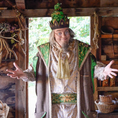 Un hombre con disfraz y corona fantasiosos posa con los brazos abiertos en una cabaña rústica en Expedition Archeon.