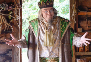 Een man in fantasierijke kostuums en kroon poseert met open armen in een rustieke hut bij Expedition Archeon.