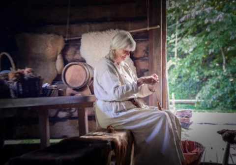 En ældre kvinde klædt i traditionelt tøj håndarbejder i en historisk hytte ved Expedition Archeon.