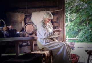 En ældre kvinde klædt i traditionelt tøj håndarbejder i en historisk hytte ved Expedition Archeon.