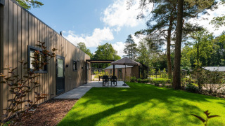 Udsigt over Loevestein Comfort lodge med moderne træfacade, grøn have og udendørs spiseplads under træer.
