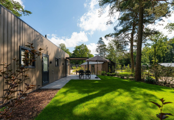 Loevestein Comfort lodge met houten buitenkant, grasveld, buitentafel en hoge bomen rondom het terrein.