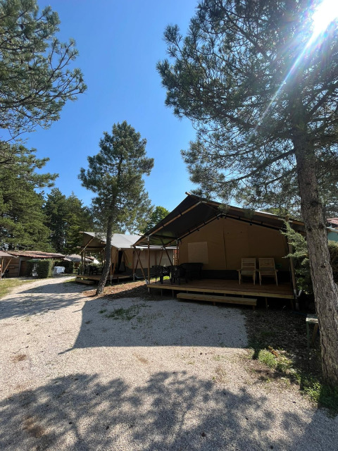 Safaritent met toilet en houten terras in een bosrijke camping, zonnige dag met blauwe hemel.