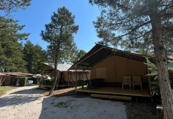 Safari telt med terrasse og toilet i et skovområde, solskin og blå himmel under trætoppene.