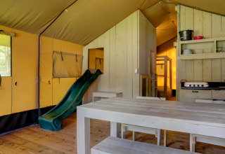 Interior de una tienda safari con mesa de madera, tobogán, cocina y literas en Familiepark Goolderheide.