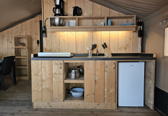 Zona de cocina en tienda safari con armarios de madera, frigorífico y utensilios en Camping Luna del Monte, Italia.