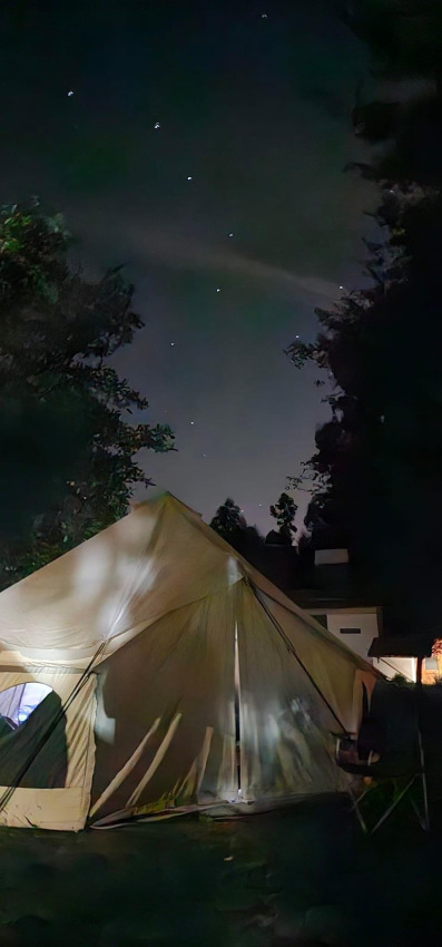 Een foto van een safaritent in het donker, omringd door bomen en een sterrijke nachtelijke hemel.
