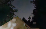 Foto di una tenda safari di notte, circondata da alberi e sotto un cielo stellato con alcune costruzioni vicine.