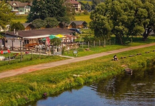 Holiday park with glamping cabins, river with canoe, and lush greenery under a clear blue sky.