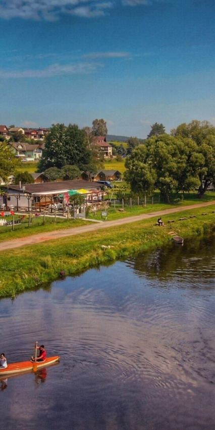 Parque vacacional con cabañas glamping, río, canoa y vegetación bajo un cielo azul claro de verano.