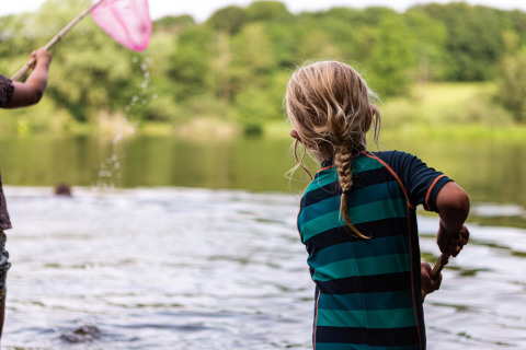 Zwei Kinder spielen am Ufer eines Sees im Vestbirk Camping, umgeben von grüner dänischer Landschaft.