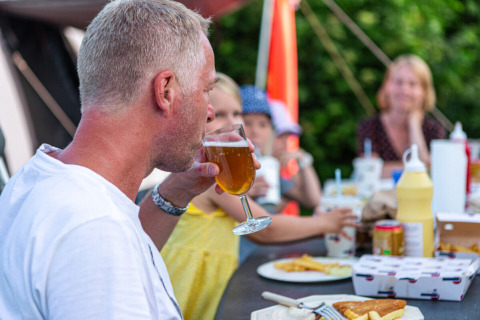 Hombre disfruta de una bebida con su familia en la mesa de Vestbirk Camping, Dinamarca, ambiente relajado.