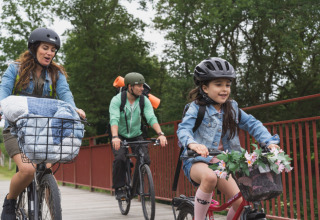 Gezin fietst met helmen over een brug bij Vestbirk Camping, Denemarken, omgeven door groene natuur.